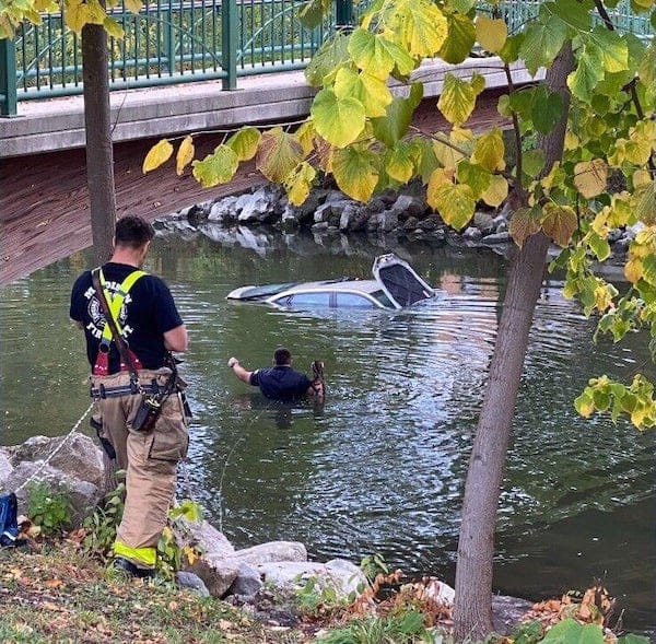 A police officer or other worker wading into the Yahara River in Madison, Wisconsin, toward a car 90 percent submerged in the water.