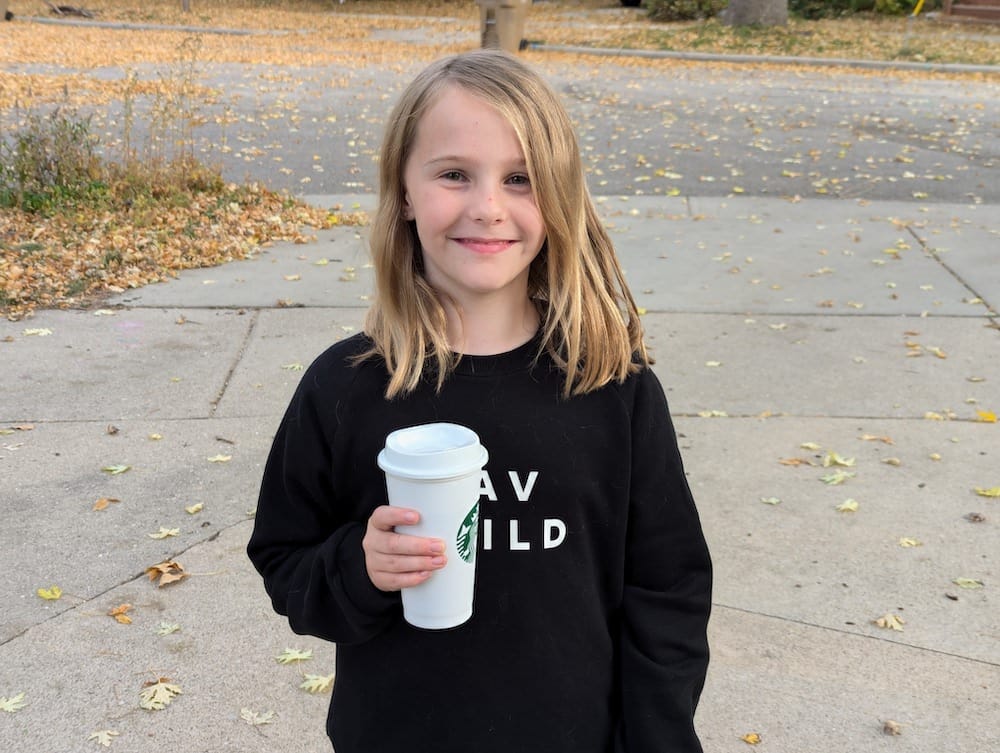 An 8-year-old child in a long-sleeved black sweatshirt reading "Fav Child," holding a reusable Starbucks cup.