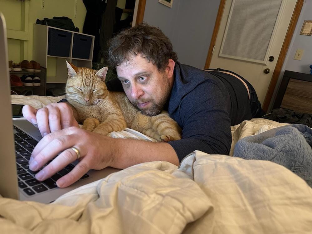 An old married man cuddles with an orange cat while he types on his laptop (the man, not the cat).