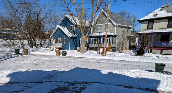 The neighborhood, snow on the ground, empty waste bins out on the street.